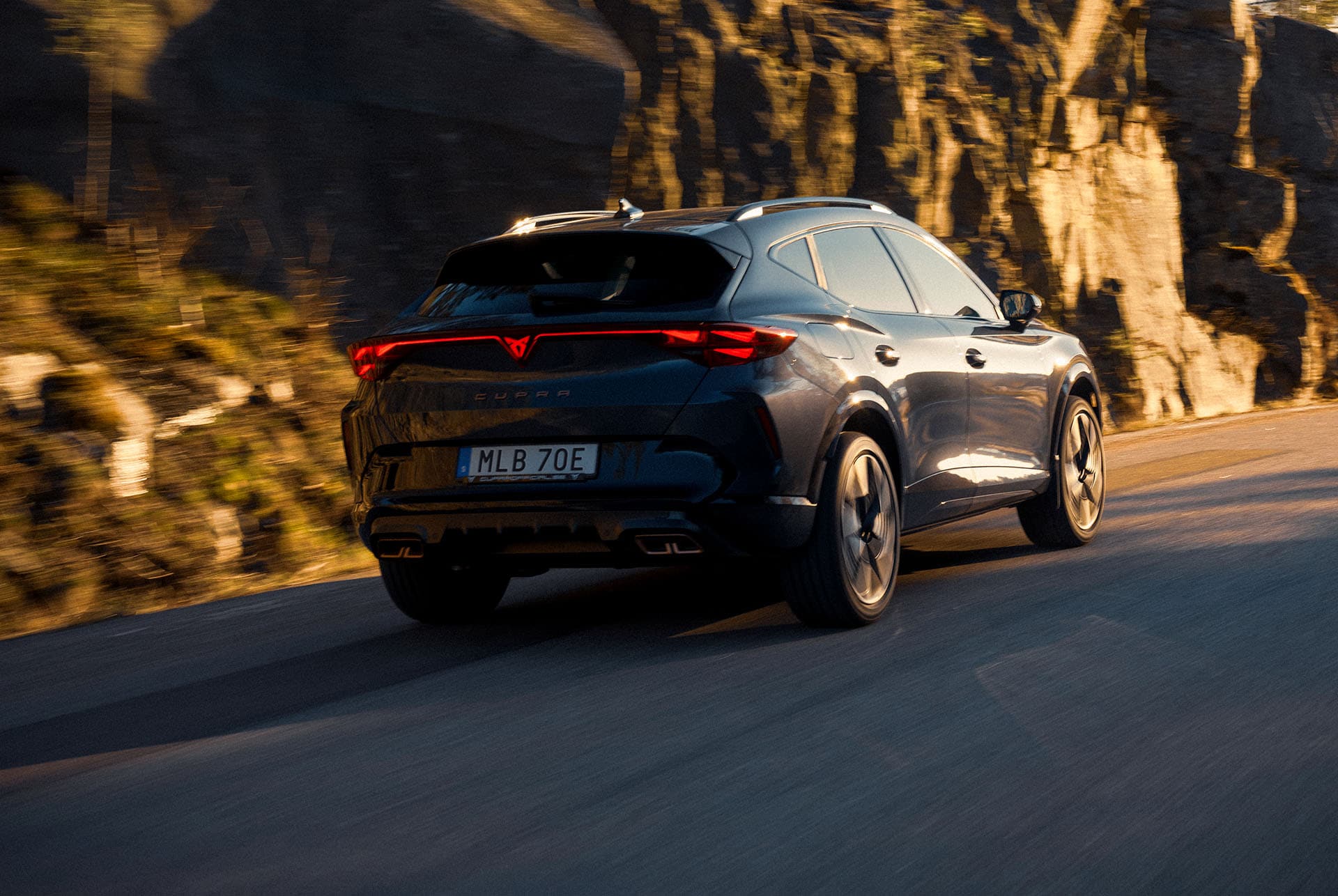 Rear three-quarter perspective of the new CUPRA Formentor hybrid car parked by waterfront and city skyline. The vehicle is angled to showcase its rear design, lights and sporty dual exhausts.
