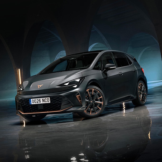 En trekvartsvy framifrån av en CUPRA Born VZ i Timanfaya Grey med koppardetaljer på hjul och grill, parkerad i ett svagt upplyst industriområde med stora stenvalv och vertikala neonljusramp.