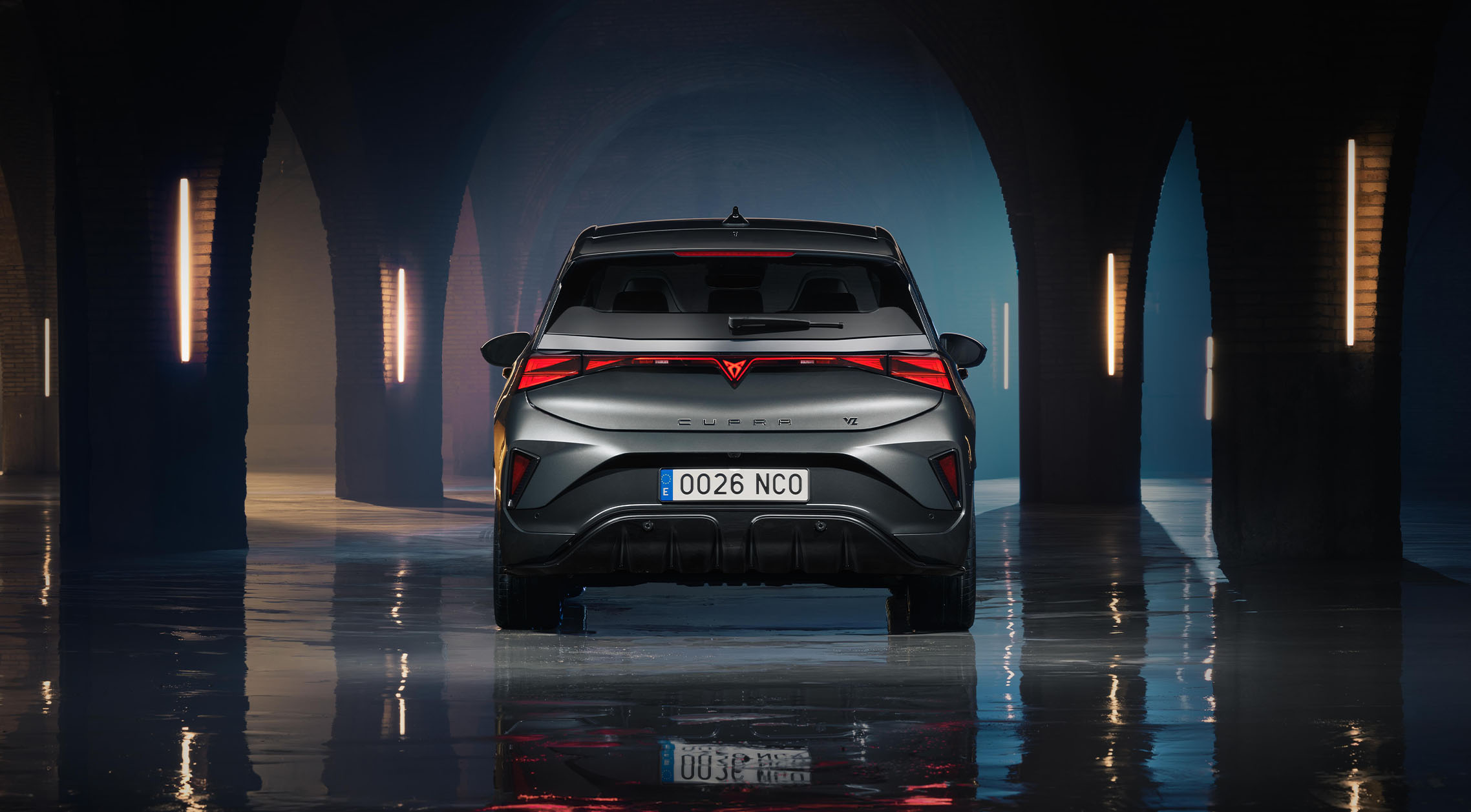A direct rear view of a CUPRA Born VZ in Timanfaya Grey, parked in a dark, vaulted industrial space with stone arches and vertical neon lighting. The image highlights the continuous LED tail light strip, the signature CUPRA logo, and the vehicle's reflection on a wet, polished concrete floor. 