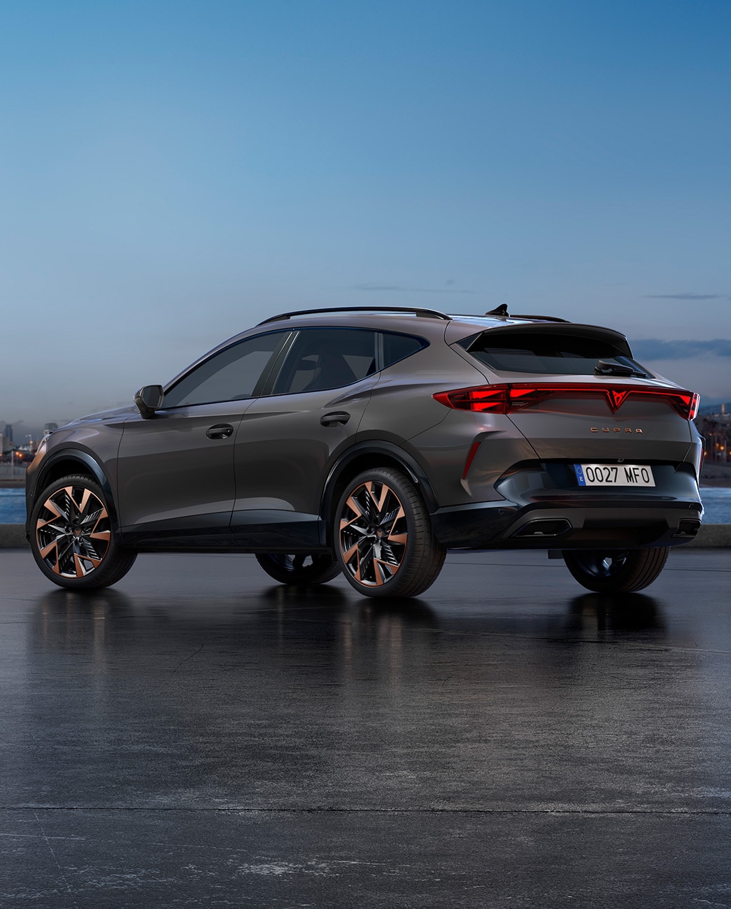 En ny CUPRA Formentor 2024 grafengrå CUV parkerad vid en strandpromenad i skymningen. I bakgrunden syns ett stadslandskap med skyskrapor och palmer upplysta av dimmande dagsljus.