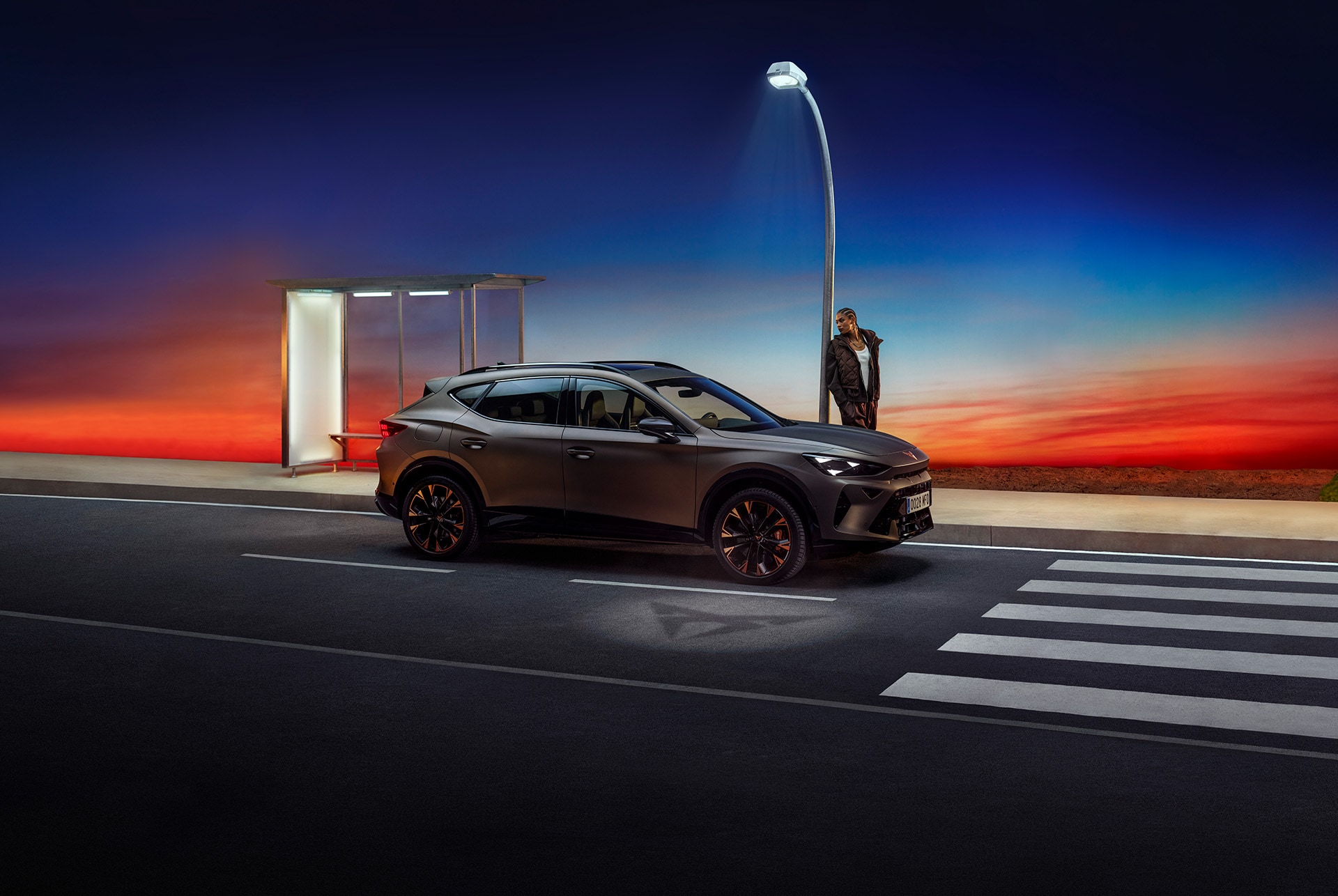 CUPRA Formentor i matt century bronze, parkerad i en byggnad mot röd himmel, laddad av en person bredvid.