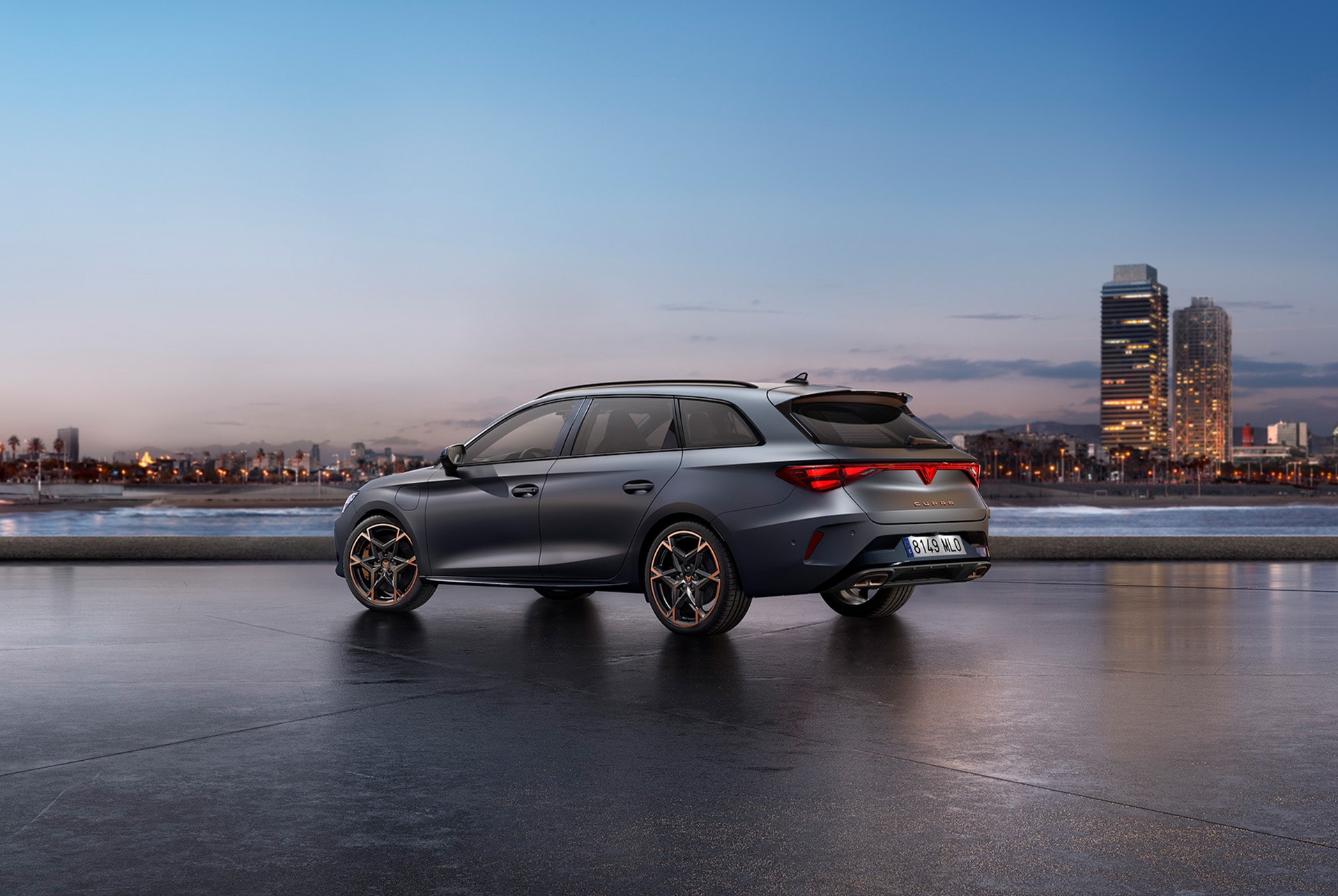 Den sportiga nya 2024 Cupra Leon Sportstourer parkerad på betong med en stadssilhuett upplyst i skymningen i bakgrunden. Bilden visar bilen i mattgrå lack.