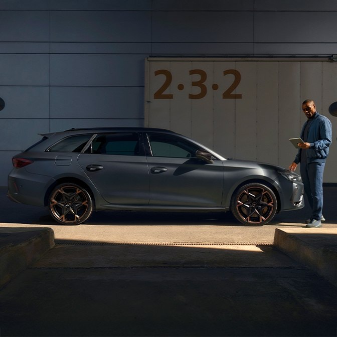 En sidoprofil av en matt grå CUPRA Leon med signaturfälgar med kopparaccent, parkerad i en betong industriell lastkaj. En man i blå jacka står längst fram i fordonet och tittar ner på en digital surfplatta medan solljuset kastar skarpa skuggor över scenen.