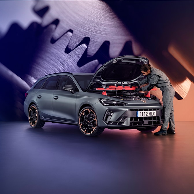 En tekniker i grå overall som inspekterar motorutrymmet på en CUPRA Terramar i en studio; bakgrunden visar en stor skärm som visar närbilder av metalliska kugghjul.