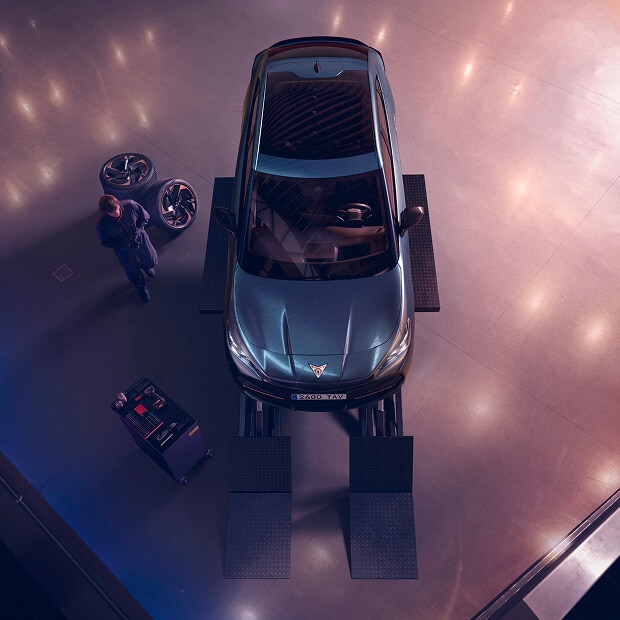 En vy ovanifrån av ett CUPRA-fordon på en hiss i ett serviceverkstad. En person syns nära några bilhjul till vänster, och en verktygsvagn finns nära bilens framsida. Golvet har subtila ljusreflektioner.